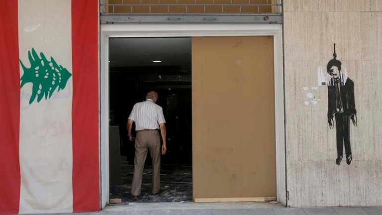 A man walks into his store completely shattered by the blast. On the wall to his left, hangs a flag of Lebanon and on the wall to his right, is a painted image of a hanged politician. Credit: Marwan Tahtah.