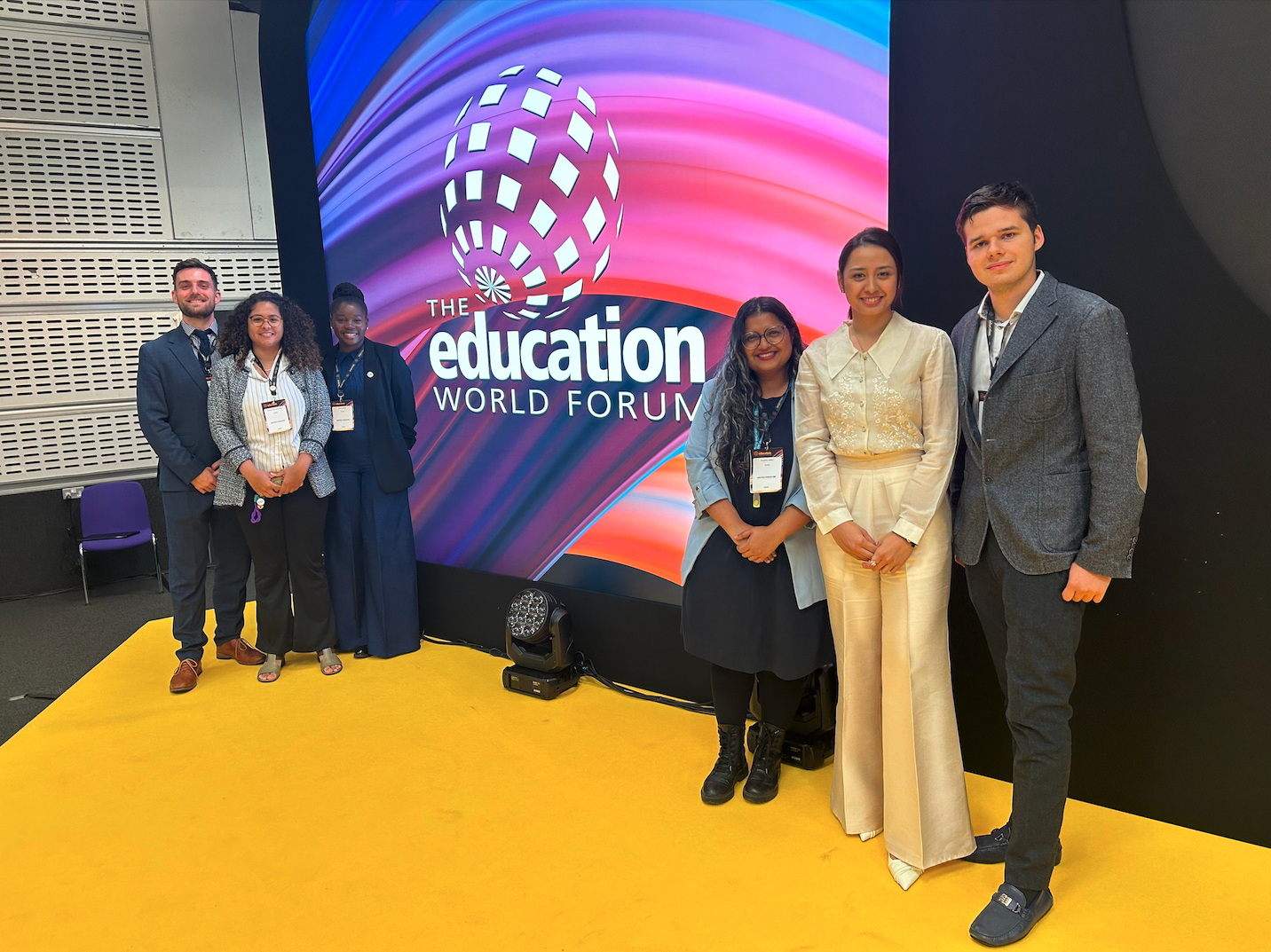 Photo of the Education World Forum logo on a screen with stripes purple and pink stripes and six smiling people in suits on either side of the logo