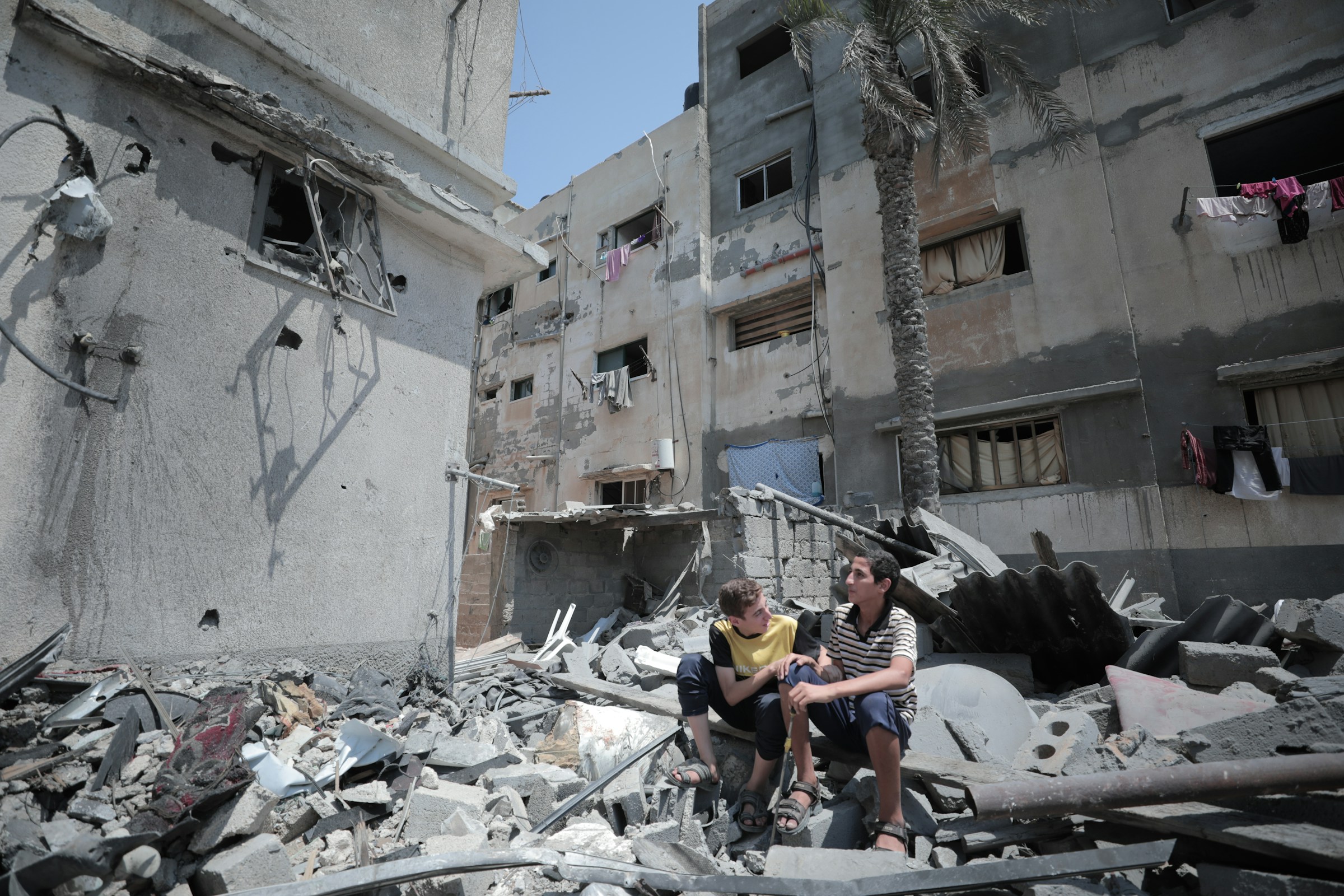 Two boys sit on rubble in Gaza, surround by buildings which have been bombed.