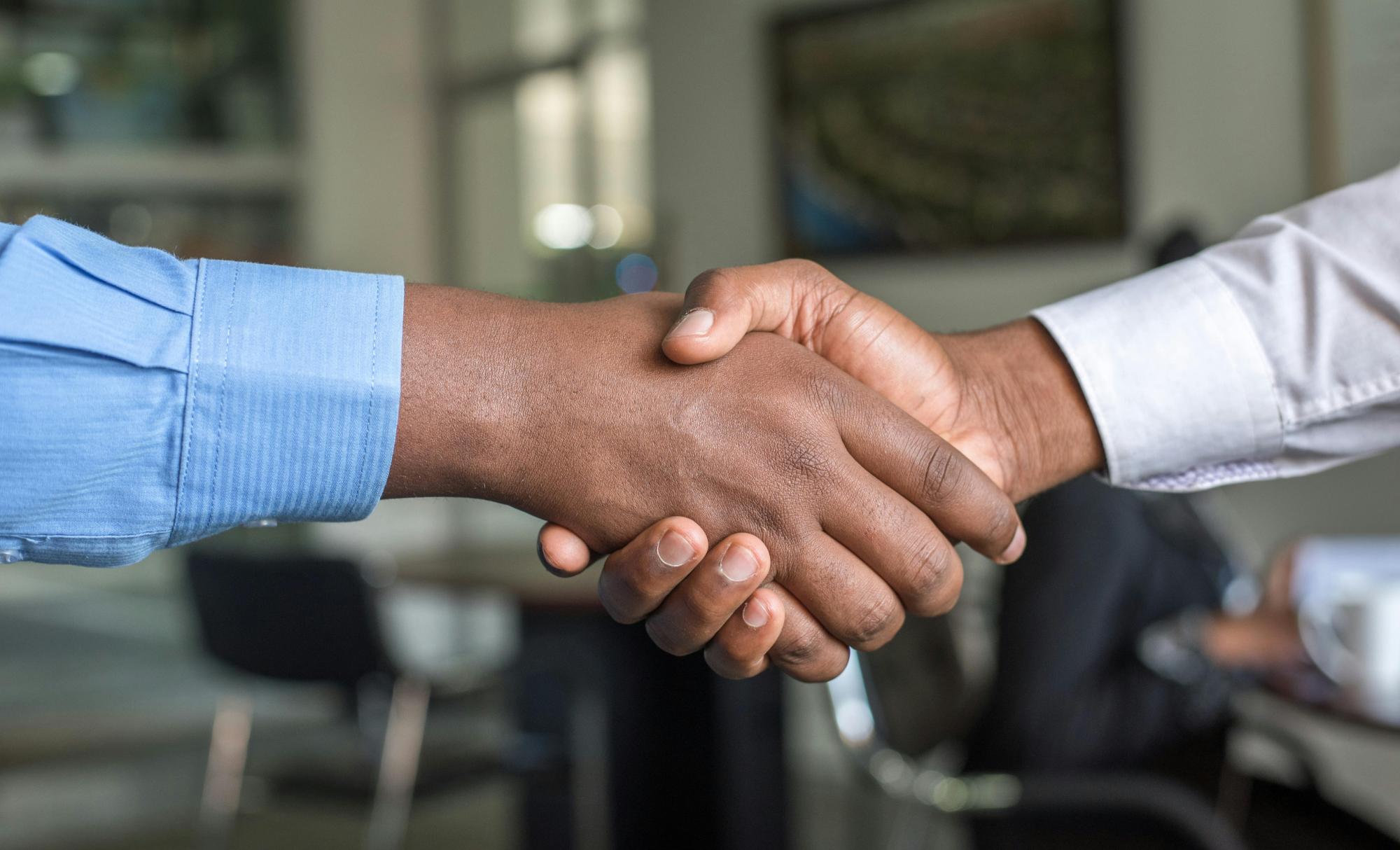 Two hands shake, both wearing shirts. An office is blurred in the background.