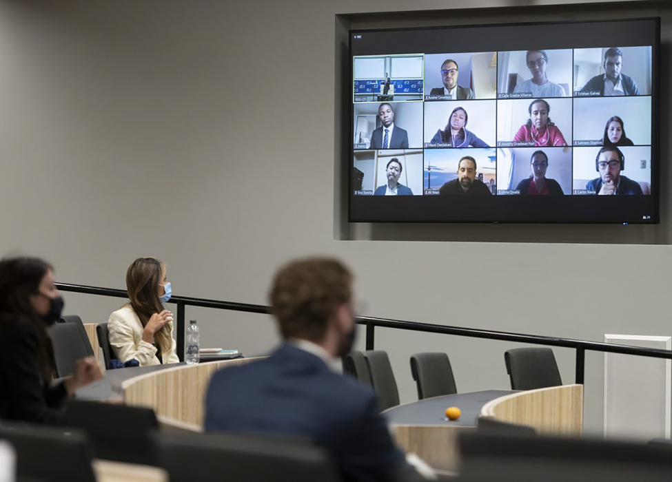 Three students sit socially distanced in a lecture theatre, facing the front where a screen displays other members of the class participating remotely